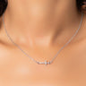 Close-up of a person wearing the NECKLACE, a delicate silver chain featuring a small horizontal cross pendant. The blurred background accentuates the contrast against the person's skin, highlighting the elegant necklace.