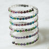 A coiled bracelet, the RING SILVER by RM Manufacturing, features a flexible silver band adorned with several rows of multicolored gemstones in shades of purple, pink, blue, and green. The bracelet is displayed on a light marble surface.