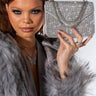 A person with sleek, styled hair and dramatic eye makeup poses with the shiny RULE THE WORLD RHINESTONE BAG held near their face. They are wearing a gray fur coat and butterfly jewelry, exuding a glamorous and fashionable vibe.