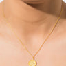 Close-up of a person wearing the SAINT CHRISTOPHER NECKLACE. The gold necklace features a small, round pendant with a textured or engraved surface. The person's skin and the necklace are prominently displayed, with minimal background visible.