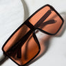 The SANTERIA OVERSIZED SUNGLASSES with large, rectangular dark red-tinted lenses and a brown frame rest on a white, textured surface. Their sleek design and modern style make them a bold fashion accessory.