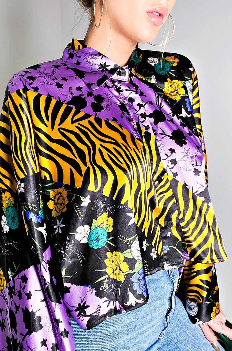 A person wearing the SECRET LIFE LONG SLEEVE BUTTON DOWN BLOUSE, adorned with eye-catching patterns that include tiger stripes, floral designs, and black silhouettes. The blouse showcases striking colors such as bold yellow, vibrant purple, lively green, and deep black. They are also dressed in a pair of blue jeans and accessorized with large hoop earrings.