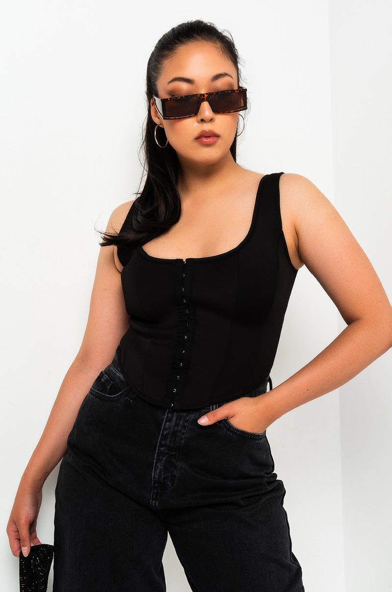 A woman stands confidently against a plain white background. She wears large, rectangular tortoiseshell sunglasses, hoop earrings, the SEXY SCUBA CORSET TOP in yellow, and high-waisted black pants. One hand rests in her pocket, and her pose is relaxed. 1