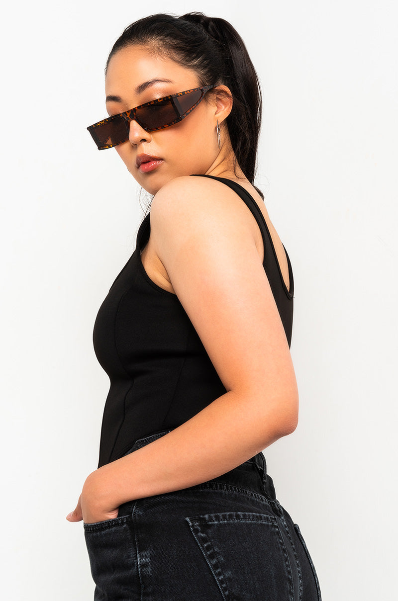 A person with long dark hair tied back, wearing rectangular tortoiseshell sunglasses, the SEXY SCUBA CORSET TOP BLACK, and black jeans stands against a white background. They have one hand resting in their pocket and are looking over their shoulder towards the camera.
