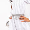 A person is dressed in a white button-down shirt with vertical pinstripes. They have the SHE'S A STAR RHINESTONE FRINGE BELT wrapped around their waist and hold a matching silver bag. The person is posing with one hand on their hip, showcasing their stylish outfit.