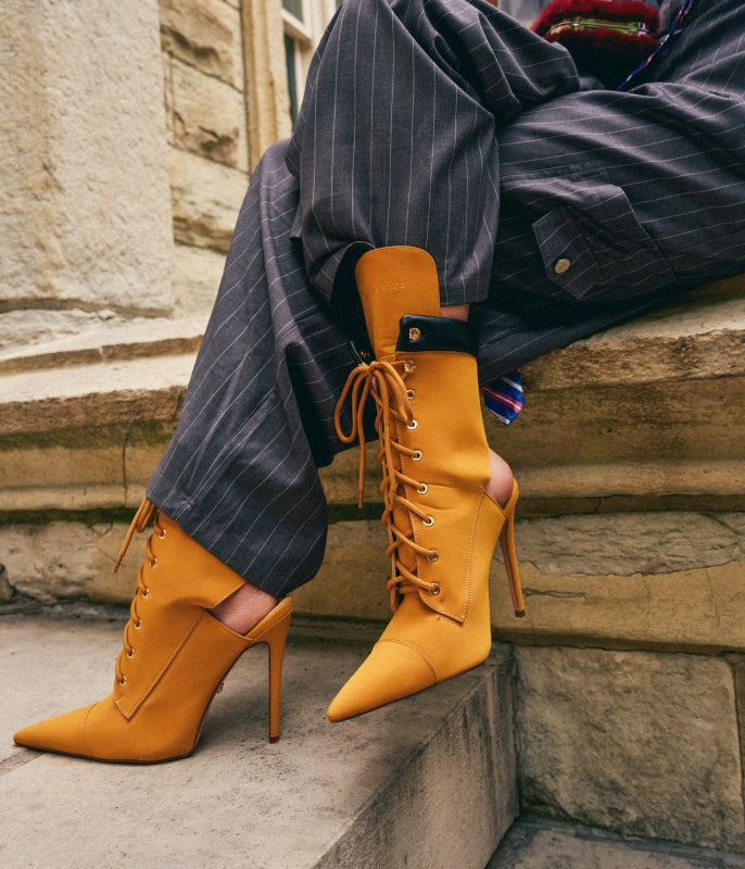 A person seated on stone steps wearing mustard yellow pointed-toe lace-up stiletto boots with slim high heels, paired with dark pinstriped trousers.