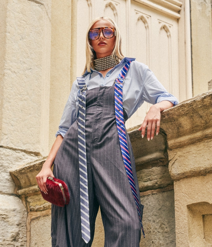 A woman stands outdoors against a stone building wearing a light blue button-up shirt, dark gray pinstriped overalls, a patterned choker, oversized blue-tinted sunglasses, and two neckties draped over the shoulders. She holds a small red clutch.
