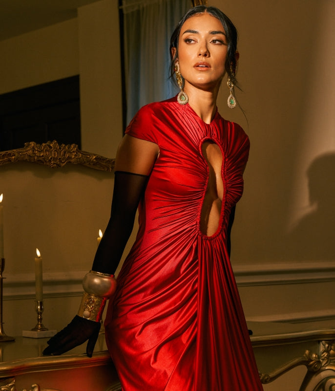 A woman wearing a dramatic red satin dress with a central cutout, black long gloves, and large dangling earrings, posed indoors near a mirror and candles.