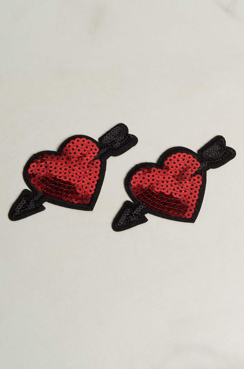 A pair of SHOT TO THE HEART VALENTINES NIPPIES, featuring embroidered red heart patches with black arrows through them and adorned with red sequins, placed against a light, neutral background.