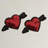 A pair of SHOT TO THE HEART VALENTINES NIPPIES, featuring embroidered red heart patches with black arrows through them and adorned with red sequins, placed against a light, neutral background.