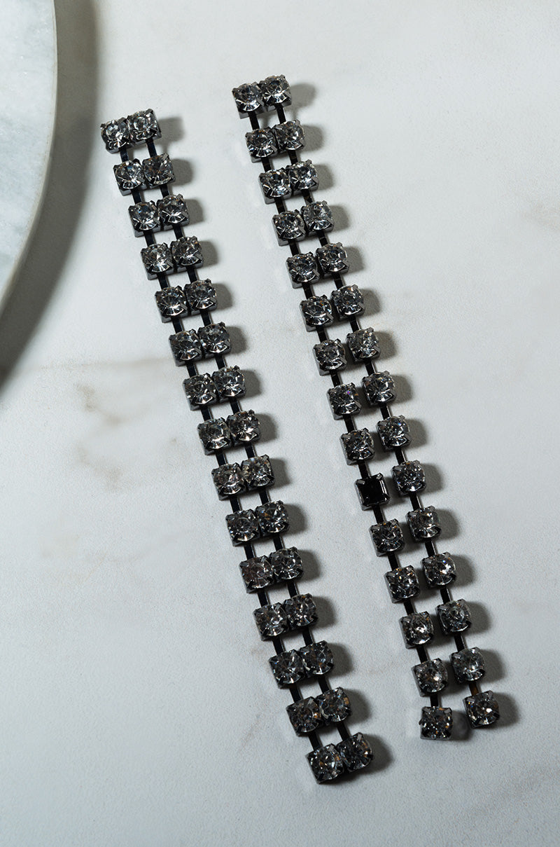 A pair of SILHOUETTE RHINESTONE DROP EARRINGS lay flat on a light-colored marble surface. These long, rectangular-shaped, silver-tone earrings feature multiple square crystal segments arranged in a grid pattern.