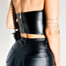 A person facing away, dressed in the MOTO DREAMER PLEATHER BANDEAU TOP, black leather pants, and a black crossbody bag with a gold chain strap. They have tattoos on their arms and wear a gold bracelet on their wrist.