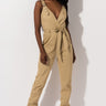 A woman poses confidently in the SLAY THE GAME JUMPSUIT by Rehab. The beige outfit boasts a deep V-neck, wide shoulder straps with buckles, a cinched waist with a fabric tie, and fitted ankle cuffs. She accessorizes with strappy high-heeled sandals and stands against a plain white background.