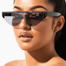 A person with sleek, dark hair pulled back tightly wears a pair of SLICK RICK RHINESTONE SUNNIES, featuring large, angular frames adorned with rhinestones along the upper edges. They have a glossy, nude lip color and a smooth complexion while posing against a plain white background.