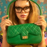 A woman with long blonde hair and cat-eye glasses holds a SLIME GREEN QUILTED PURSE in front of her face. She is wearing a colorful, patterned outfit and is set against a background with floral designs.