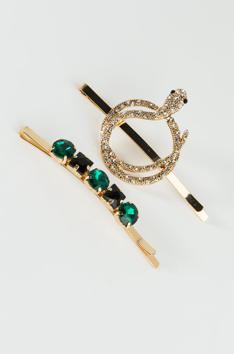 Two stylish hairpins from the SNAKEY RHINESTONE HAIR PINS collection are placed on a white surface. One hairpin is adorned with a snake design encrusted with clear rhinestones, while the other features a linear arrangement of alternating green and black gemstones on a gold base.