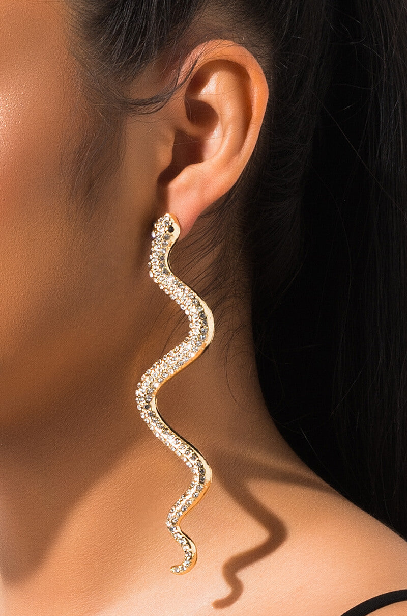 Close-up of a person's ear adorned with the SNEAKY SNAKE EARRINGS GOLD, a large, gold snake-shaped earring encrusted with sparkling rhinestones. The earring extends significantly below the earlobe, curving gracefully to mimic the undulating shape of a snake.