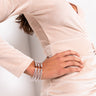 A woman in a light beige, form-fitting, long-sleeve dress stands with one hand on her hip. She is wearing a multi-strand gold bracelet and silver dangling earrings. Her dark wavy hair falls over her shoulders, and the overall look is stylish and elegant.