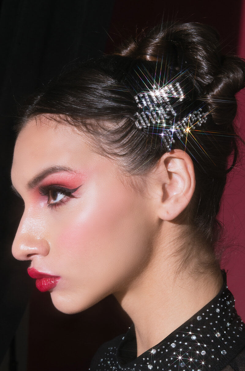 A close-up side profile of a person with intricate makeup, featuring bold red eyeshadow, pink blush, and red lipstick. Their hair is styled in an elegant updo, adorned with the SPICY PRINCESS HAIR CLIP SET's glimmering star-shaped hairpiece. The background is a blend of dark and pink hues.