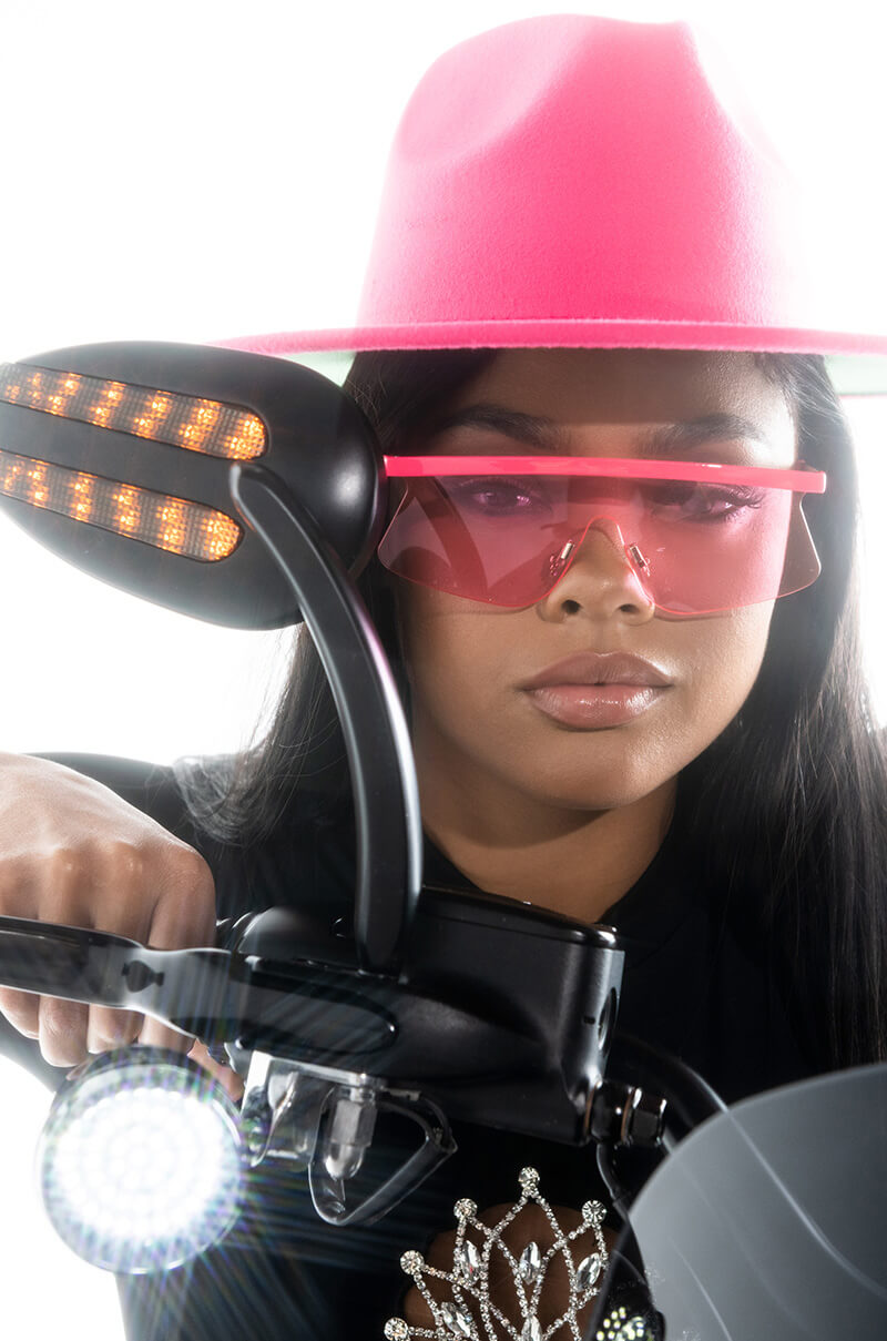 A person wearing a bright pink hat and YAPPER TREND SUNNIES rides a motorcycle. The motorcycle's rearview mirror and headlight are visible. The person has long, dark hair and a serious expression. The image is taken from a low-angle, close-up perspective.
