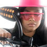 A person wearing a bright pink hat and YAPPER TREND SUNNIES rides a motorcycle. The motorcycle's rearview mirror and headlight are visible. The person has long, dark hair and a serious expression. The image is taken from a low-angle, close-up perspective.