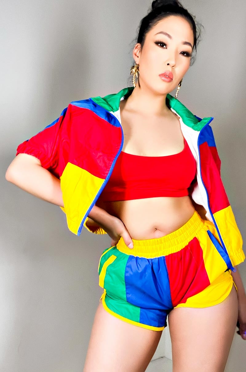 A woman is posing against a plain background wearing a colorful outfit. She has a confident expression and is dressed in a red sports bra and the NUMBER ONE FAN FULL ZIP SHORT SLEEVE JACKET paired with matching shorts in red, yellow, blue, and green blocks.