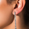 Close-up of a person's ear wearing the BELLA EARRING SET. The earrings feature small, sparkling gemstones arranged in a row, cascading down to end with a larger, round gemstone. The indoor setting with warm lighting enhances the intricate details of the jewelry.