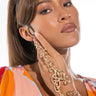 A woman with long brown hair in a high ponytail poses with her hand near her face. She is wearing a colorful, patterned top and the SUMMERTIME MAGIC HAND BRACELET, which wraps around her fingers and wrist in ornate golden swirls. She has a neutral, serious expression.