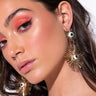 A woman with long, wavy brown hair and vibrant coral eye makeup gazes at the camera. She is wearing striking SUN IN MY EYES DANGLE EARRINGS, which are gold fan-shaped pieces adorned with blue and white evil eye symbols. The lighting highlights her glowing skin and the details of her makeup and jewelry.