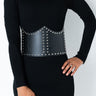 A person wearing a black ribbed turtleneck dress with long sleeves. They have a hand resting on their hip and are accessorized with the SUPER FREAK STUDDED CORSET BELT, a wide black leather belt adorned with metal studs. Their curly hair is partially visible, and they are standing against a plain background.