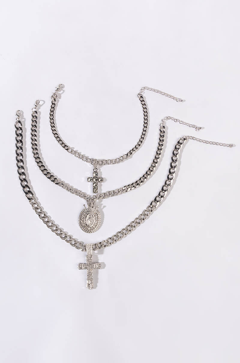 The HOLY WOMAN LAYERED RHINESTONE NECKLACE SET IN SILVER features three silver-toned chunky chain necklaces on a light background, each with a distinct pendant: a circular medallion, a rhinestone-encrusted religious pave charm, and a larger cross, merging style with spiritual elegance.