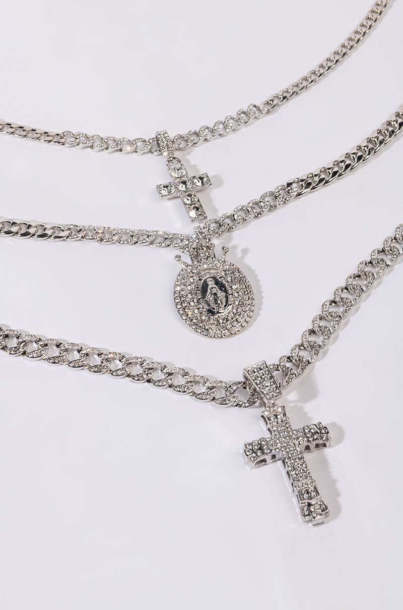 The HOLY WOMAN LAYERED RHINESTONE NECKLACE SET IN SILVER features three layered necklaces on a white background. The top and bottom ones display ornate cross pendants, while the middle resembles a rhinestone chain with a detailed round medallion, all adorned with sparkling stones that catch the light beautifully.