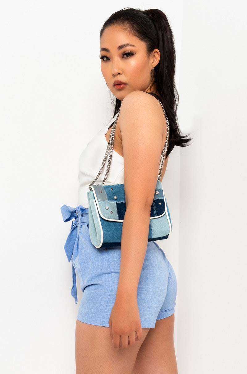 A woman in the SWEET SUMMER SPAGHETTI STRAP ROMPER stands against a white background. She has a blue handbag with metal studs slung over her shoulder and is looking back over her shoulder towards the camera.

