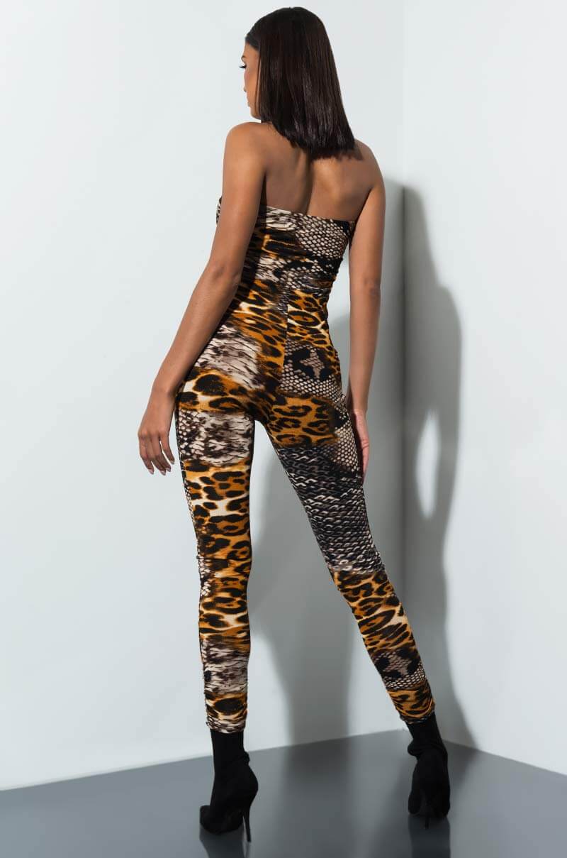 A person with shoulder-length dark hair stands in a corner, facing away. They are wearing the "TAKE ME ON A VACAY PLAYSUIT," a strapless bodysuit showcasing an eye-catching mixed animal print pattern and black boots. The playsuit features patterns reminiscent of leopard and snake prints.