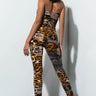 A person with shoulder-length dark hair stands in a corner, facing away. They are wearing the "TAKE ME ON A VACAY PLAYSUIT," a strapless bodysuit showcasing an eye-catching mixed animal print pattern and black boots. The playsuit features patterns reminiscent of leopard and snake prints.