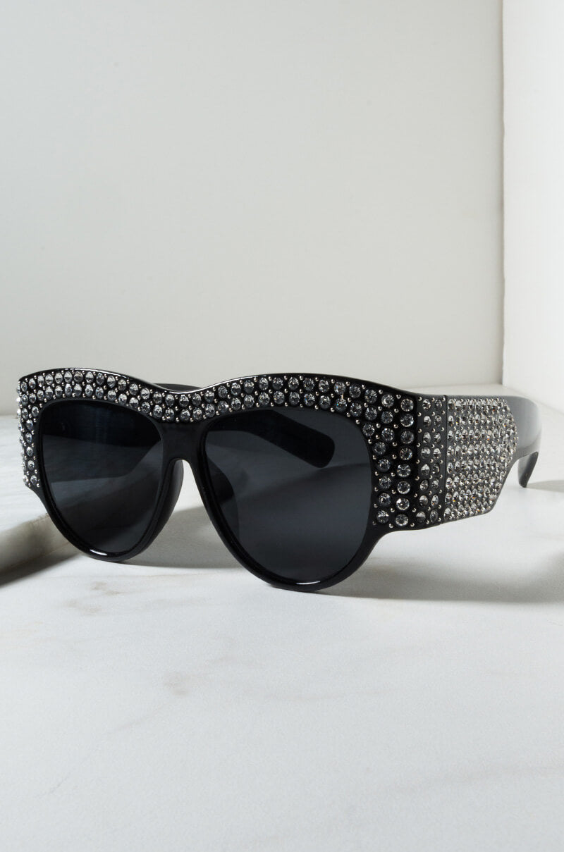 TELL ME NOTHING RHINESTONE SUNGLASSES ASSORTED feature an array of small, shimmering rhinestones along the top rim and temples of their black oversized frame. The dark-tinted lenses enhance the bold, glamorous style. These stunning sunglasses are showcased on a light-colored surface with a minimalist background.