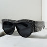 The TELL ME NOTHING RHINESTONE SUNGLASSES BLACK SILVER feature a pair of black oversized sunglasses with dark lenses. The frame is embellished with numerous small, sparkling rhinestones that cover the top and sides, creating a glamorous and eye-catching design. The background is a simple, light-colored surface.