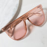 The THANK YOU NEXT OVERSIZED SUNGLASSES are a pair of large, translucent peach-colored sunglasses with a sleek, modern design featuring rivet details at the temples. They rest on a smooth, light marble surface.
