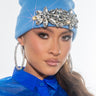 A person wearing THE AFTER PARTY GEM KNIT HAT, a blue beanie adorned with large, sparkling rhinestones, and oversized hoop earrings. They are also dressed in a sheer, bright blue top and are gazing intently at the camera against a plain, light-colored background.