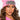 A person with long, wavy brown hair is wearing THE AFTER PARTY GEM KNIT HAT, a bright pink beanie adorned with a large, jeweled brooch. They have soft makeup featuring bold eyeliner and nude lipstick, while the background is plain white.