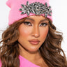 A person with long, wavy brown hair is wearing THE AFTER PARTY GEM KNIT HAT, a bright pink beanie adorned with a large, jeweled brooch. They have soft makeup featuring bold eyeliner and nude lipstick, while the background is plain white.