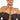 A person with braided hair is wearing an off-the-shoulder black top and THE BEST IS YET TO COME BODY CHAIN draped over their shoulders. The background is plain and bright, emphasizing the detailed design of the body chain. The person gazes confidently at the camera.