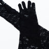 A pair of THE MET GALA GLOVE in black lace are laid flat on a white surface, with one glove partially overlapping the other. The delicate, intricate floral lace pattern is clearly visible, emphasizing the gloves' elegant design.