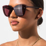 A person with sleek, dark hair styled in a low bun is wearing the TIERRA OVERSIZED SUNGLASSES in a large, square, dark red design. They have a gold nose ring and small hoop earrings, along with multiple layered gold necklaces. The background is plain and white.