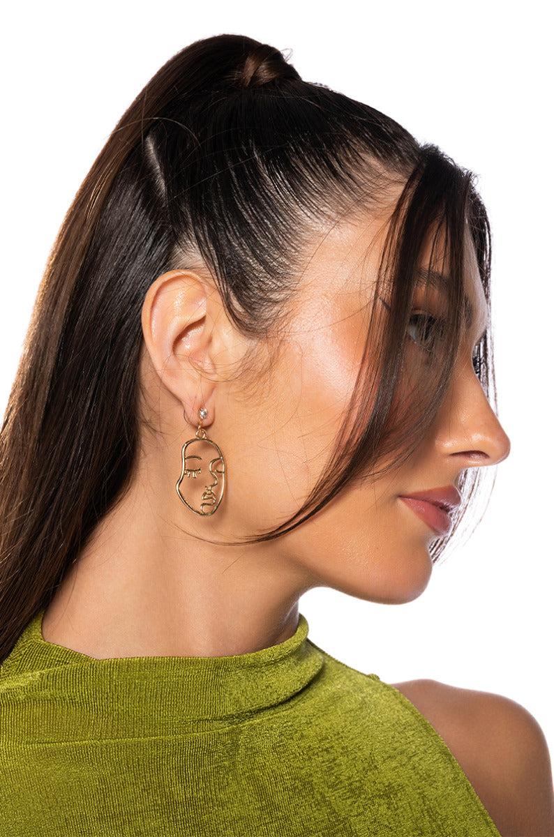 A woman with long, dark hair styled back in a ponytail is shown in profile. She is wearing a green sleeveless top and stunning FACE IT GOLD EARRINGS shaped like faux gold facial silhouettes. Her skin is glowing, and her hair gently frames part of her face against the white background.