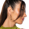 A woman with long, dark hair styled back in a ponytail is shown in profile. She is wearing a green sleeveless top and stunning FACE IT GOLD EARRINGS shaped like faux gold facial silhouettes. Her skin is glowing, and her hair gently frames part of her face against the white background.