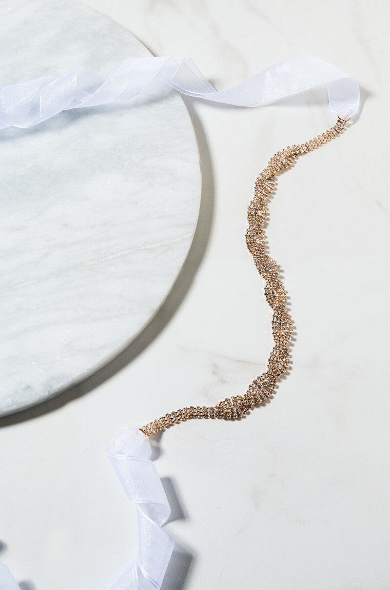 The TO THE STARS TIE BACK BELT is laid out elegantly on a white marble surface. Positioned diagonally, the belt features a white, semi-transparent ribbon woven through its links, adding a soft decorative touch.