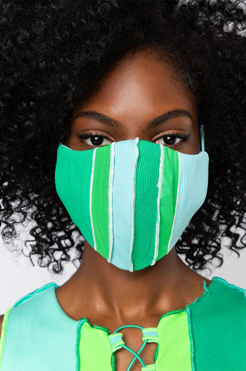 A person with curly black hair wears the TOO GOOD TO BE TRUE FASHION FACE COVER, which features vertical green and blue stripes. They are also dressed in a green and blue top that has lace-up detail at the neckline. The background is plain and light-colored.