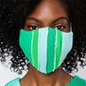 A person with curly black hair wears the TOO GOOD TO BE TRUE FASHION FACE COVER, which features vertical green and blue stripes. They are also dressed in a green and blue top that has lace-up detail at the neckline. The background is plain and light-colored.