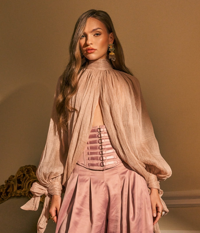 A woman modeling a dusty pink high-neck sheer blouse with voluminous sleeves and a matching satin corset-style waist detail, paired with pleated pink bottoms, against a warm beige wall.